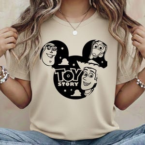 K&ouml;nnte beinhalten: Beiges T-Shirt mit schwarzer Grafik der Toy Story-Charaktere in einer Mickey-Mouse-Silhouette. Der Text "TOY STORY" befindet sich unter den Figuren. Das Shirt besteht aus einem weichen Material.