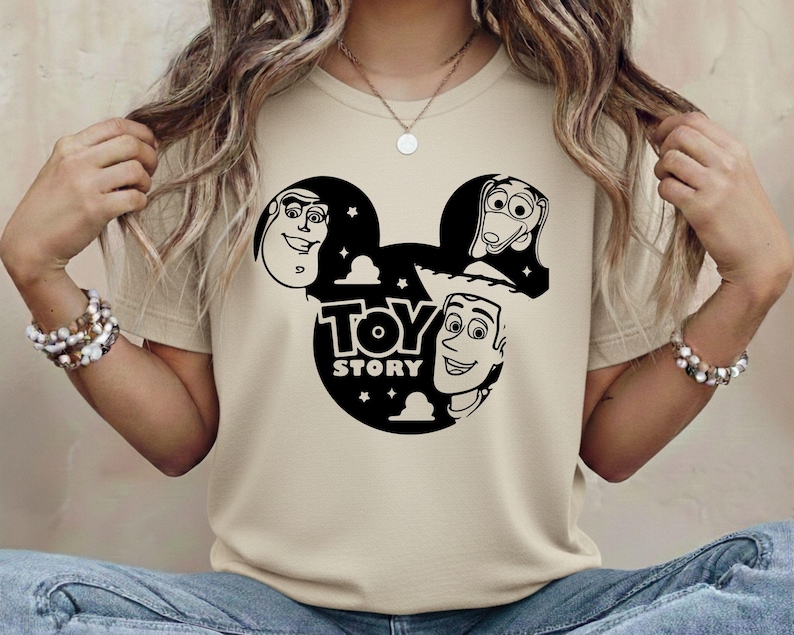 K&ouml;nnte beinhalten: Beiges T-Shirt mit schwarzer Grafik der Toy Story-Charaktere in einer Mickey-Mouse-Silhouette. Der Text "TOY STORY" befindet sich unter den Figuren. Das Shirt besteht aus einem weichen Material.
