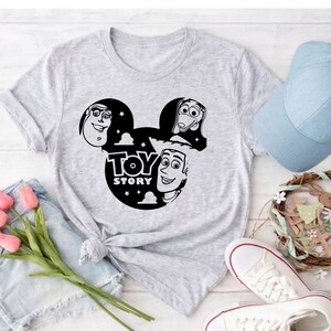 K&ouml;nnte beinhalten: Hellgraues T-Shirt mit einer schwarz-wei&szlig;en Grafik der Toy Story-Charaktere in einem von Mickey Mouse inspirierten Design, einschlie&szlig;lich des Textes "TOY STORY". Eine hellblaue Baseballkappe, wei&szlig;e Turnschuhe und ein dekorativer Kranz sind ebenfalls zu sehen.