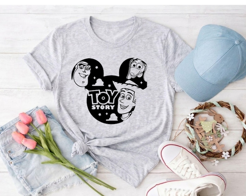 K&ouml;nnte beinhalten: Hellgraues T-Shirt mit einer schwarz-wei&szlig;en Grafik der Toy Story-Charaktere in einem von Mickey Mouse inspirierten Design, einschlie&szlig;lich des Textes "TOY STORY". Eine hellblaue Baseballkappe, wei&szlig;e Turnschuhe und ein dekorativer Kranz sind ebenfalls zu sehen.