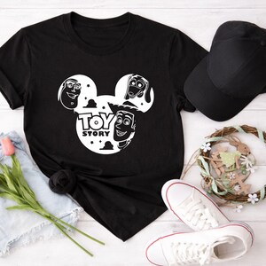 K&ouml;nnte beinhalten: Schwarzes T-Shirt mit einem wei&szlig;en Aufdruck von Toy Story-Charakteren in einem von Mickey Mouse inspirierten Design. Eine schwarze Baseballkappe, wei&szlig;e Turnschuhe mit roter Umrandung und ein dekorativer Kranz sind ebenfalls zu sehen.