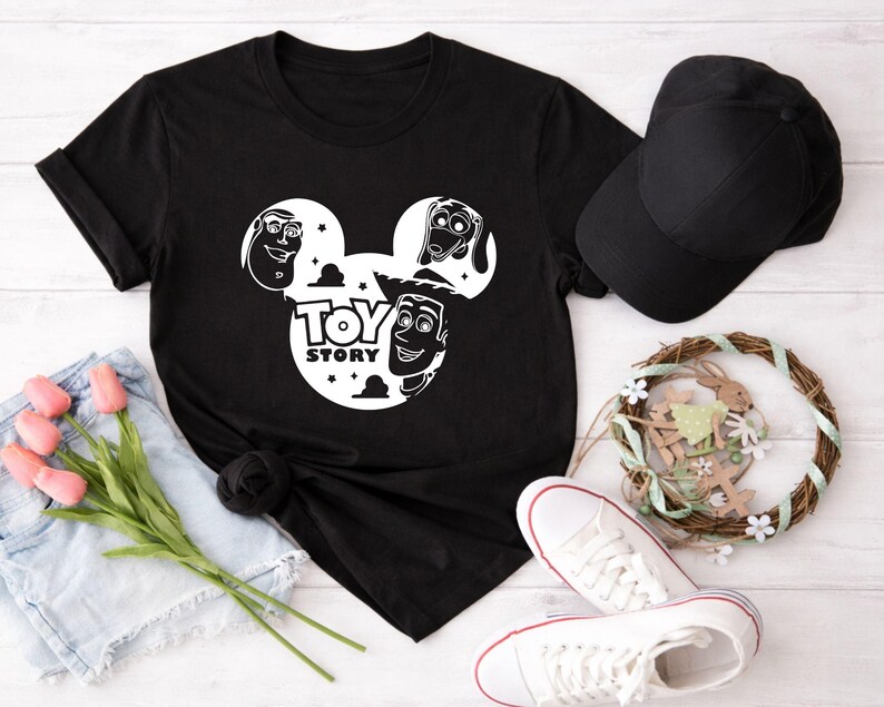 K&ouml;nnte beinhalten: Schwarzes T-Shirt mit einem wei&szlig;en Aufdruck von Toy Story-Charakteren in einem von Mickey Mouse inspirierten Design. Eine schwarze Baseballkappe, wei&szlig;e Turnschuhe mit roter Umrandung und ein dekorativer Kranz sind ebenfalls zu sehen.