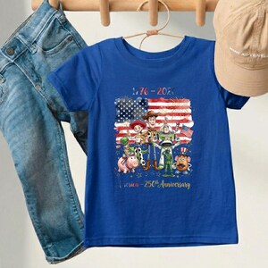 May include: A blue t-shirt featuring a graphic of animated characters in front of an American flag. The shirt has the text "1776-2026" and "America - 250th Anniversary". A pair of blue jeans and a tan baseball cap are also in the image.
