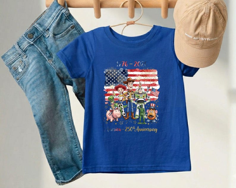 May include: A blue t-shirt featuring a graphic of animated characters in front of an American flag. The shirt has the text "1776-2026" and "America - 250th Anniversary". A pair of blue jeans and a tan baseball cap are also in the image.