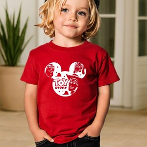 K&ouml;nnte beinhalten: Ein rotes T-Shirt mit einem wei&szlig;en Aufdruck des Toy Story-Logos und Charaktergesichtern. Das kurz&auml;rmelige Shirt besteht aus einem weichen Material. Das Design zeigt die Gesichter von Woody, Buzz Lightyear und anderen Charakteren.
