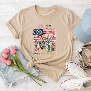 May include: Beige t-shirt featuring a graphic of cartoon characters in front of an American flag. The text reads "1776-2026" and "America - 250th Anniversary". A blue baseball cap, white sneakers, and denim shorts are also visible.