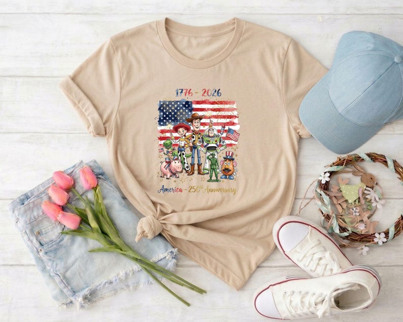 May include: Beige t-shirt featuring a graphic of cartoon characters in front of an American flag. The text reads "1776-2026" and "America - 250th Anniversary". A blue baseball cap, white sneakers, and denim shorts are also visible.