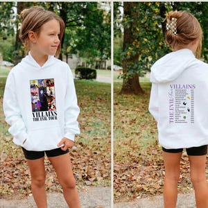Op de afbeelding: Witte hoodie met een afbeelding op de voorkant met verschillende tekenfilm-schurken en de tekst "Villains The Evil Tour". De achterkant van de hoodie heeft een lijst met schurken. Het model draagt een zwarte short.