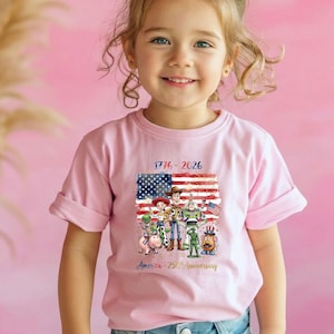 May include: A light pink t-shirt featuring a graphic of the American flag and cartoon characters. The shirt has the text "1776-2026" and "America - 250th Anniversary". The shirt is being worn by a young child.