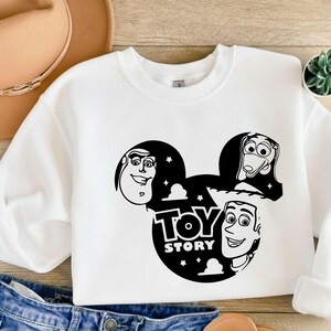 K&ouml;nnte beinhalten: Wei&szlig;es Sweatshirt mit Rundhalsausschnitt und schwarzer Grafik der Toy Story-Charaktere Buzz Lightyear, Slinky Dog und Woody in einem von Mickey Mouse inspirierten Design. Die Worte "Toy Story" sind ebenfalls in der Grafik enthalten.