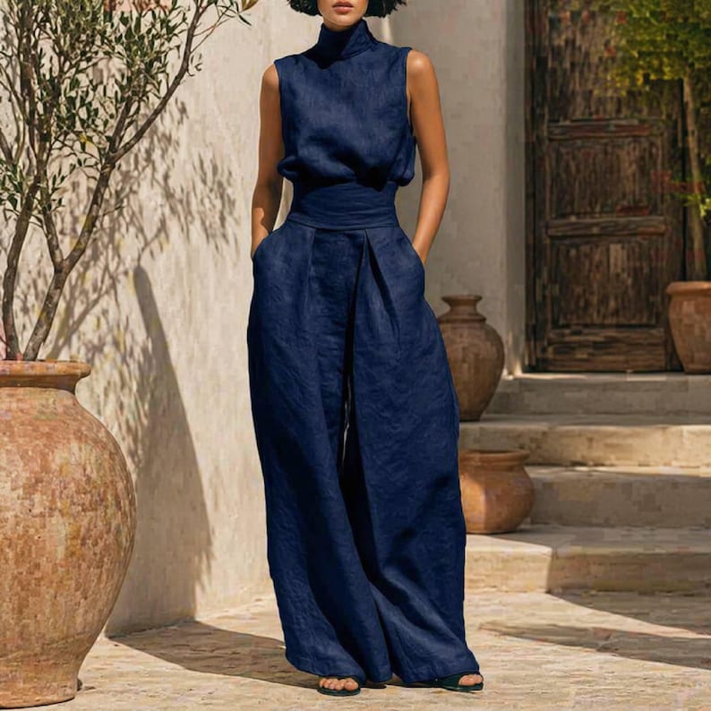 May include: A sleeveless, navy blue jumpsuit with a high neckline and wide-leg pants. The jumpsuit features a cinched waist and pockets. The model is standing outdoors near a stone wall and wooden door.
