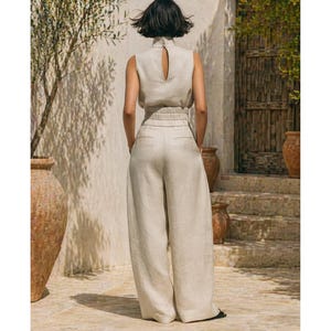 May include: A beige linen two-piece outfit. The sleeveless top has a high neckline with a keyhole back. The high-waisted, wide-leg pants have a relaxed fit. The outfit is made of a lightweight fabric, perfect for warm weather.