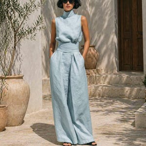 May include: A light blue sleeveless jumpsuit with a high neckline and wide-leg pants. The jumpsuit features a cinched waist and is made of a lightweight fabric. The model is wearing sunglasses. The outfit is suitable for warm weather.