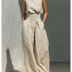 May include: A sleeveless, light beige linen jumpsuit. The top has a high neckline and a fitted waist. The wide-leg pants have pockets. The outfit is made of a lightweight, natural fabric, perfect for warm weather. The model is wearing the outfit.