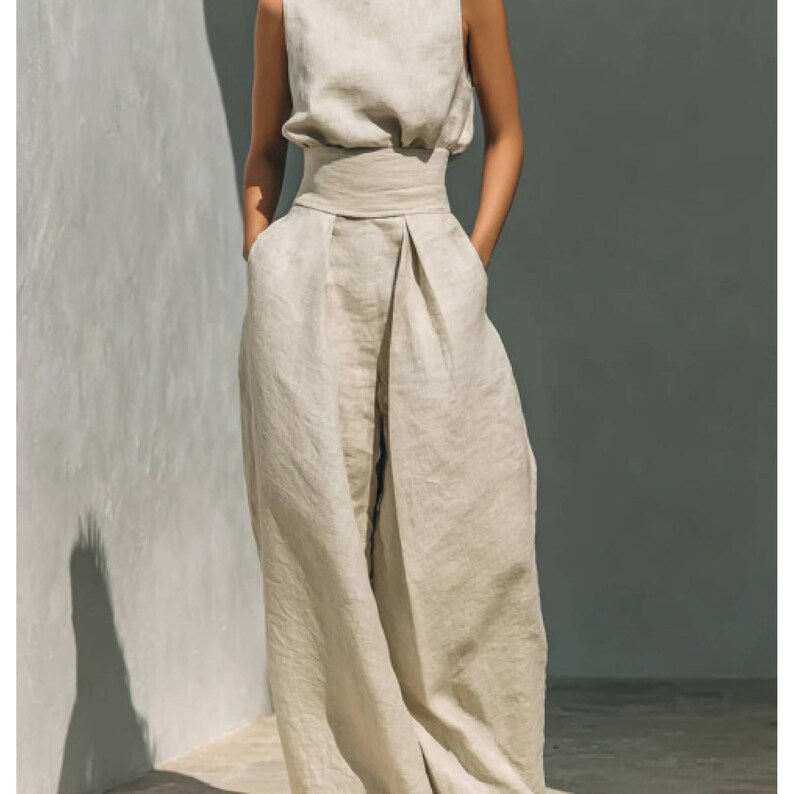 May include: A sleeveless, light beige linen jumpsuit. The top has a high neckline and a fitted waist. The wide-leg pants have pockets. The outfit is made of a lightweight, natural fabric, perfect for warm weather. The model is wearing the outfit.