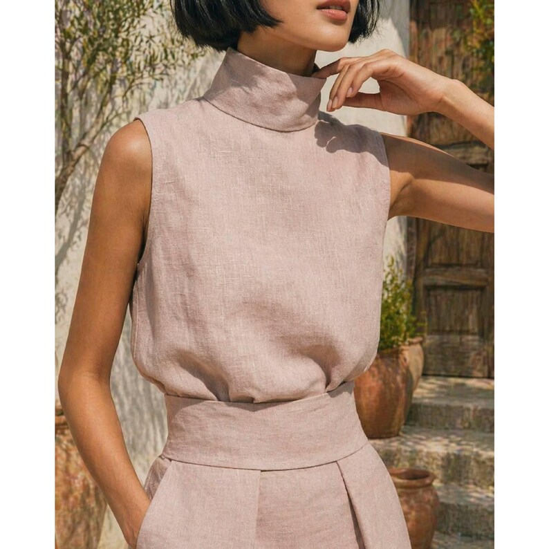 May include: A sleeveless, blush-colored top with a high neckline and a matching wide waistband. The top is made of a textured fabric, possibly linen, and is paired with high-waisted pants. The outfit is simple and elegant.