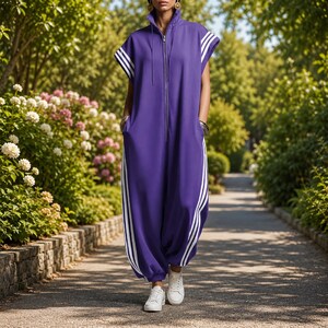 Puede incluir: Mono morado con rayas blancas en los laterales y en las mangas. El mono tiene una cremallera en la parte delantera y bolsillos. La persona lleva zapatillas blancas. El atuendo se usa al aire libre en una pasarela pavimentada.