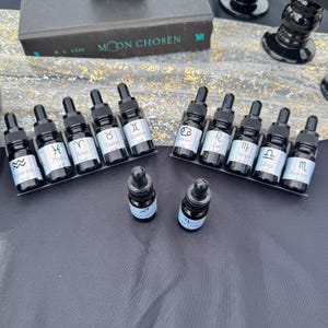 May include: A collection of twelve small dark glass dropper bottles, each featuring a white label with a zodiac sign symbol and name. The bottles are arranged in two rows of five on black stands, with two additional bottles placed in the front.