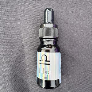 May include: A small dark glass dropper bottle with a black cap. The label features a holographic finish, the Libra zodiac symbol, and the text "Libra 10 ml". The bottle is designed for dispensing liquid contents through the integrated glass dropper.
