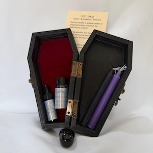 May include: A black wooden coffin-shaped box with a red velvet interior, containing two small glass bottles labeled GOTHMAS and Silent Night, two taper candles in black and purple, and a small black obsidian stone placed in front.