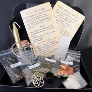 May include: A Yule spell jar kit in a black box containing a gold candle, a small glass jar with a cork, a silver pentacle charm, and clear plastic bags labeled Tiger's Eye, Rosemary, Cedar, Orange Peel, and Salt, with two instruction cards.