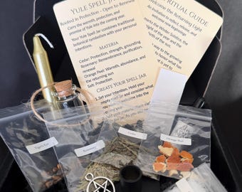 May include: A Yule spell jar kit in a black box containing a gold candle, a small glass jar with a cork, a silver pentacle charm, and clear plastic bags labelled Tiger's Eye, Rosemary, Cedar, Orange Peel, and Salt, with two instruction cards.
