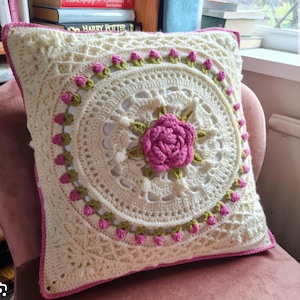 Op de afbeelding: Crèmekleurig, vierkant gehaakt kussen met een centrale roze roos en een rand van roze knoppen. Het kussen heeft ingewikkeld openwerk en een roze rand, waardoor een handgemaakt, bloemenontwerp ontstaat. Het kussen ligt op een roze stoel.