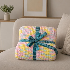 Könnte beinhalten: Eine gefaltete Decke in Pastelltönen von Rosa, Gelb und Blau, gebunden mit einem türkisfarbenen Band. Die Decke besteht aus einem weichen, strukturierten Material und wird auf einem neutral getönten Sofa präsentiert. Ca. 100 cm x 150 cm.