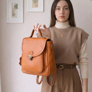 Könnte beinhalten: Ein beigefarbener Lederrucksack mit Tragegriff, einem Frontklappenverschluss mit Metallschnalle und verstellbaren Schultergurten. Die Tasche hat eine genarbte Textur und eine strukturierte rechteckige Form mit einer Vordertasche unter der Klappe.