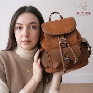 Puede incluir: Una mochila de ante y cuero marrón con una solapa superior con cierre de hebilla, un bolsillo frontal con hebilla y dos bolsillos laterales. La bolsa tiene un asa superior y costuras visibles. El logotipo CLUTCHKIN BAG COLLECTION es visible.