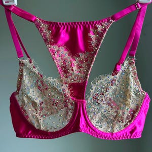 May include: A vibrant hot pink underwire bra featuring beige lace overlays on the cups and center panel. The lace is embellished with numerous small, iridescent rhinestones that catch the light. It includes adjustable pink straps and a racerback.