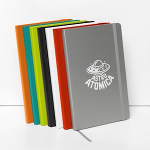 Peut inclure: Une rangée de six carnets à couverture rigide disposés en éventail. Les couleurs de l'arrière vers l'avant sont orange, bleu sarcelle, vert citron, noir, blanc, rouge et un carnet gris avec un motif d'OVNI blanc et le texte ASTRO ATOMICA.