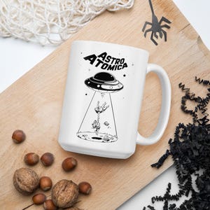 May include: A white ceramic mug featuring a black graphic illustration of a UFO beaming up a person. The text ASTRO ATOMICA is printed above the flying saucer. The design includes stars, a small landscape, and a laptop on the ground below the beam.