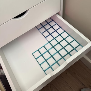 May include: A white drawer unit with an open drawer containing a blue and teal grid-style plastic organiser insert. The organiser features a square pattern and is positioned in the corner of the drawer, leaving a portion of the white base visible.