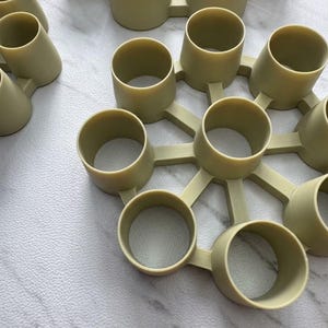 May include: A light olive green plastic organizer featuring a central circular hub connected to eight evenly spaced cylindrical holders. The structure is rigid with smooth, matte surfaces and open tops, designed for holding multiple small items.