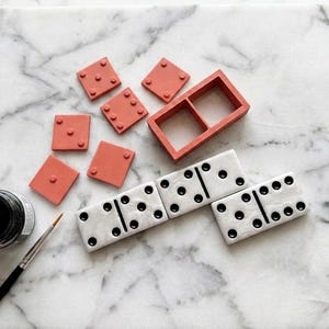 May include: A set of dominoes including three finished white tiles with black pips, several unfinished terracotta-colored square tiles, a rectangular terracotta frame, a small jar of black paint, and a thin paintbrush for crafting.