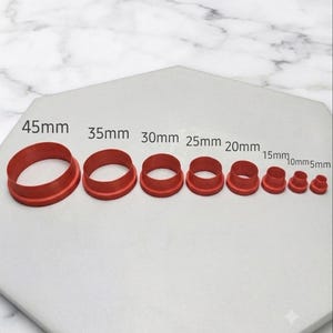 May include: A set of eight circular red cutters arranged in descending order of size, labeled with measurements from 45mm down to 5mm. Each cutter features a wide base and a raised cylindrical rim, designed for precise cutting tasks.