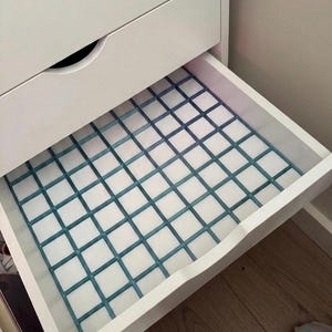 May include: A white drawer organizer featuring a blue grid pattern insert. The organizer is placed inside an open white drawer of a cabinet. The grid structure creates multiple square compartments for storage, providing a clear view of the drawer base.