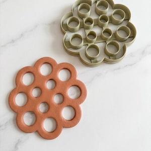 May include: A sage green plastic cookie cutter with a multi-circular floral shape and a corresponding terracotta-colored clay cutout featuring a matching pattern of various sized circular holes arranged in a cluster.