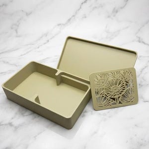 May include: A light olive green rectangular storage box with an open lid and a separate rectangular lid insert featuring a detailed laser-cut floral pattern. The box has a smooth finish and internal dividers, providing a structured container.