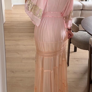 Pu&ograve; includere: Un abito lungo rosa chiaro con vita smock, maniche ampie con ricami floreali dorati e una gonna a balze con una fascia orizzontale in pizzo all'uncinetto. Il capo arriva fino a terra e presenta una silhouette morbida e fluida.
