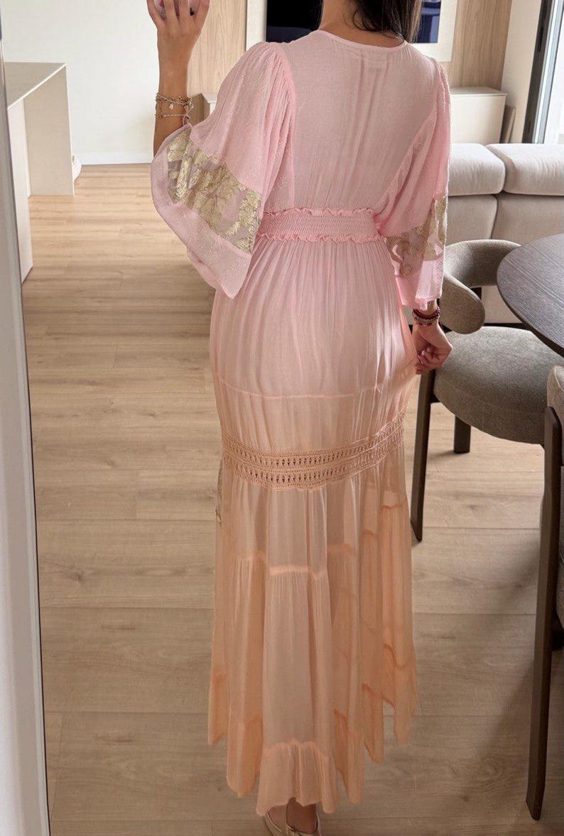 Pu&ograve; includere: Un abito lungo rosa chiaro con vita smock, maniche ampie con ricami floreali dorati e una gonna a balze con una fascia orizzontale in pizzo all'uncinetto. Il capo arriva fino a terra e presenta una silhouette morbida e fluida.