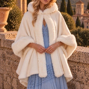 May include: A cream-coloured hooded cape featuring a thick faux fur trim along the edges and a decorative silver clasp at the neckline, worn over a light blue tiered dress with intricate lace detailing on the bodice and skirt panels.