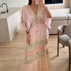 Pu&ograve; includere: Un abito lungo color rosa chiaro con un profondo scollo a V impreziosito da paillettes dorate, maniche ampie e una vita con coulisse e nappe rosa. La gonna presenta pannelli orizzontali di ricami floreali dorati e inserti in pizzo.