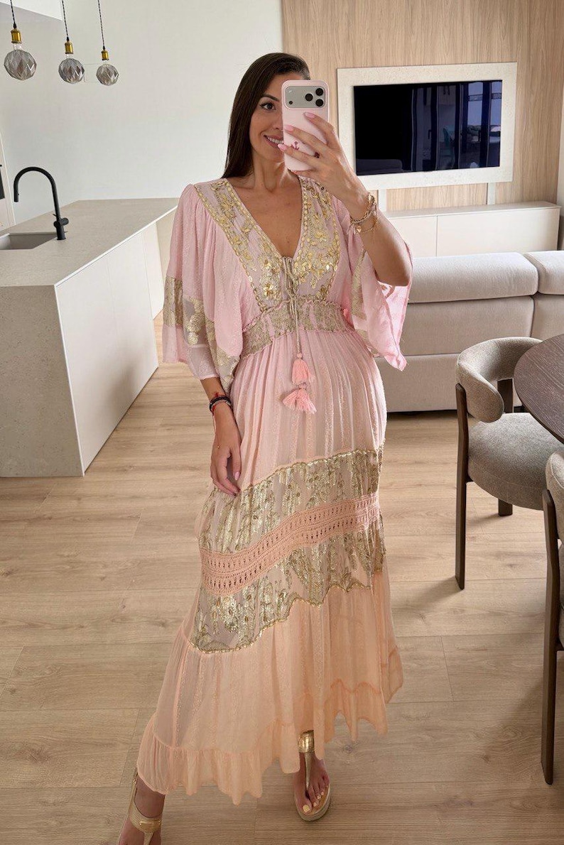 Pu&ograve; includere: Un abito lungo color rosa chiaro con un profondo scollo a V impreziosito da paillettes dorate, maniche ampie e una vita con coulisse e nappe rosa. La gonna presenta pannelli orizzontali di ricami floreali dorati e inserti in pizzo.