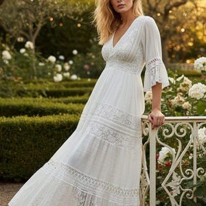 Può includere: Un lungo abito bianco con scollo a V e dettagli in pizzo. L'abito presenta una gonna a balze e maniche svasate. La donna è in un giardino con rose bianche e una recinzione decorativa. L'abito è realizzato in tessuto leggero.