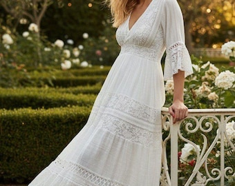 May include: A white, floor-length dress with a V-neck and lace detailing. The dress features a tiered skirt and bell sleeves. The woman is standing in a garden with white roses and a decorative fence. The dress is made of a lightweight fabric.