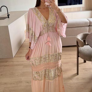 Pu&ograve; includere: Un abito lungo rosa chiaro con profondo scollo a V, ampie maniche a volant e vita con coulisse e due nappe. Il capo &egrave; decorato con motivi floreali in oro metallizzato, dettagli in pizzo e una gonna fluida a balze.