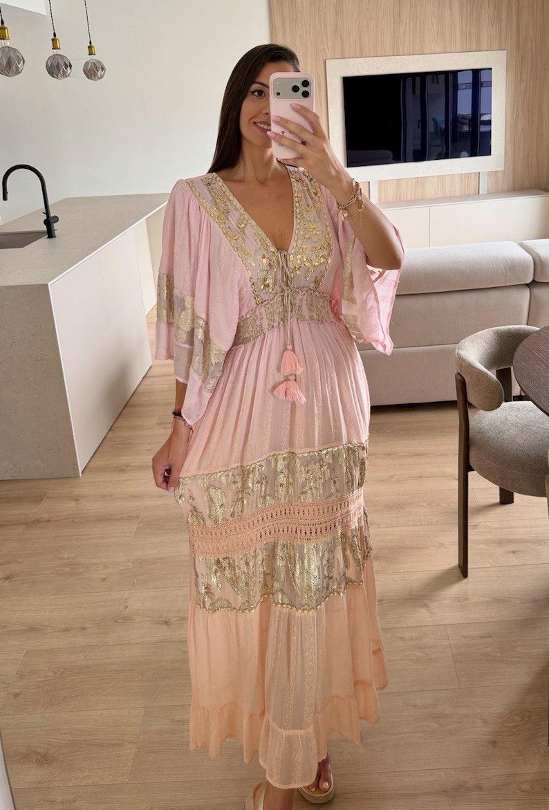 Pu&ograve; includere: Un abito lungo rosa chiaro con profondo scollo a V, ampie maniche a volant e vita con coulisse e due nappe. Il capo &egrave; decorato con motivi floreali in oro metallizzato, dettagli in pizzo e una gonna fluida a balze.