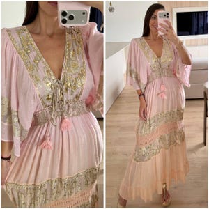 Pu&ograve; includere: Un abito lungo rosa chiaro con profondo scollo a V impreziosito da ricami metallici dorati, maniche ampie e vita arricciata. La gonna presenta fasce orizzontali in tessuto metallico dorato, dettagli in pizzo e un orlo con volant.